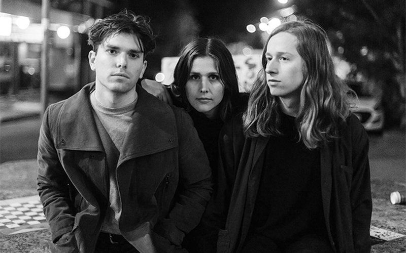 Drei junge Erwachsene posieren ernst vor nächtlicher Stadtkulisse, schwarz-weiß.